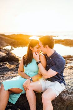 a man and woman are sitting on rocks near the water, one is biting into the other's mouth