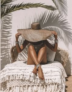 a woman sitting on top of a bed under a palm tree wearing a straw hat