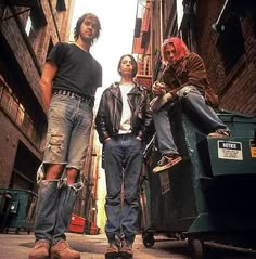 three young men standing in an alleyway next to a dumpster and garbage can