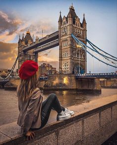 a woman is sitting on the edge of a wall looking at the tower bridge in london