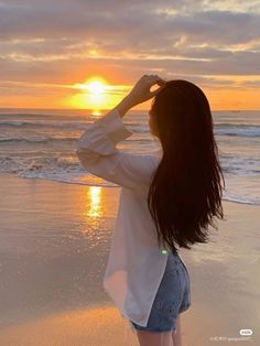 a woman standing on top of a sandy beach next to the ocean at sunset with her hair blowing in the wind