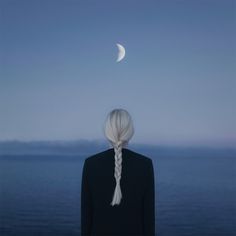 a woman with long white hair standing in front of the ocean looking at the moon