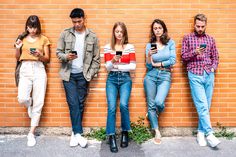 four people leaning against a brick wall looking at their cell phones while one person checks his phone