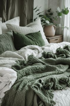 an unmade bed with green blankets and pillows on top of it, in front of a potted plant