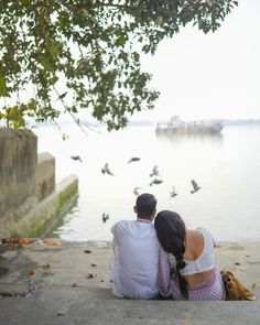 a man and woman sitting on the ground next to water with birds flying around them