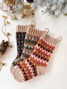 two crocheted christmas stockings hanging from hooks on a white table with pine cones