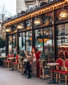 two women sitting at tables in front of a restaurant with red chairs and lights on the windows