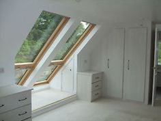 an attic bedroom with slanted windows and white furniture in the foreground is a dresser, chest of drawers, and closets