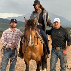 two men and a woman standing next to a brown horse on a dirt field with mountains in the background