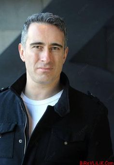 a man wearing a black jacket and white t - shirt is looking at the camera