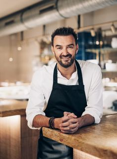 a man in an apron leaning on a counter with his arms crossed and smiling at the camera