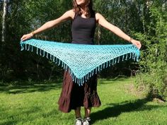 a woman standing in the grass holding up a blue crocheted shawl