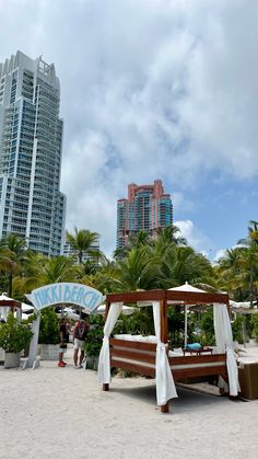 a wooden bed sitting on top of a sandy beach next to tall buildings and palm trees