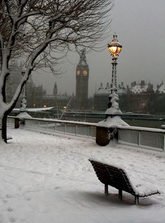 a park bench covered in snow next to a lamp post and clock tower on a snowy day