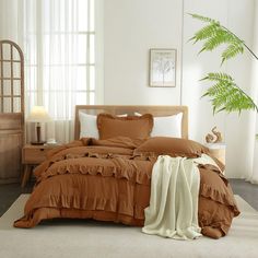 a bed with brown sheets and pillows in a white room next to a large green plant