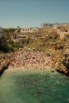 many people are swimming in the water near some cliffs and buildings, while others stand on the beach