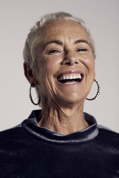 an older woman with white hair and large hoop earrings smiles at the camera while wearing a black top