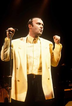 a man in a yellow jacket and black pants holding a microphone on stage with his right hand up