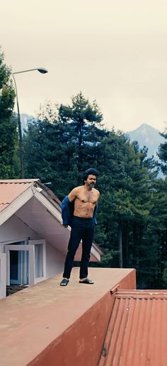 a shirtless man standing on top of a roof next to a building with trees in the background