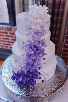 a three tiered cake with purple flowers on the top and bottom, sitting on a silver platter