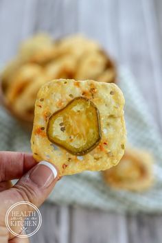 a hand holding up a cracker with a pickle on it and other snacks in the background