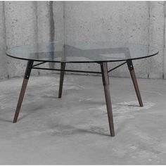 an oval glass table with wooden legs in front of a concrete wall and cement floor