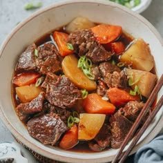 a bowl filled with beef and carrots next to chopsticks on a table