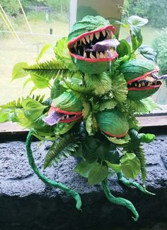 a fake plant with green leaves and monster teeth on it's head, in front of a window