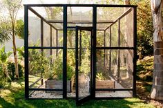 a glass house with plants inside in the grass near trees and bushes, on a sunny day