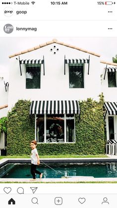 a little boy standing in front of a pool next to a white house with black and white striped awnings