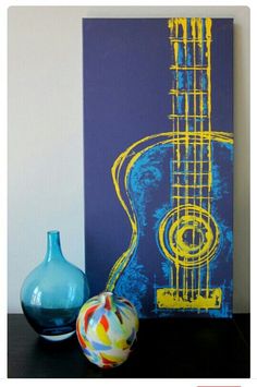 a blue vase sitting next to a painting and a guitar on a wall behind it