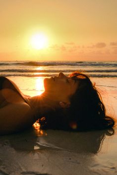 a woman laying on top of a sandy beach next to the ocean at sun set