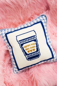 a pink furry surface with a blue and white cross stitch pillow on it that says happy you are here