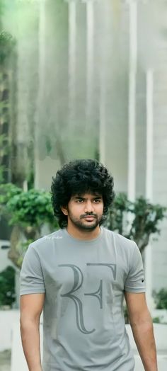 a man with curly black hair standing in front of a building wearing a t - shirt