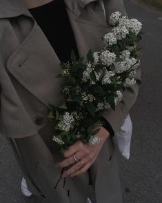 a woman in a trench coat holding a bouquet of white flowers on her left hand