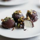 three chocolate covered desserts on a white plate with nuts and other food items in the background