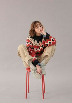 a young woman sitting on top of a red stool wearing a sweater and pants, with her hand under her chin