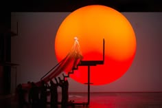 people are standing in front of a giant orange ball with a woman on it's back