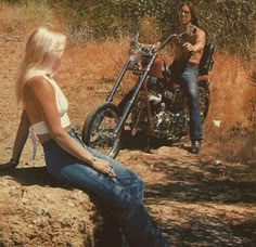 a woman sitting on the ground next to a motorcycle and another man standing behind her