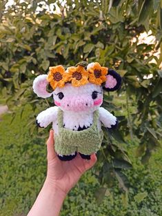 a hand holding a small stuffed cow with flowers on it's head in front of some bushes