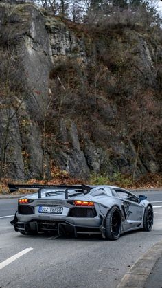 a grey sports car driving down the road in front of a rock wall and trees