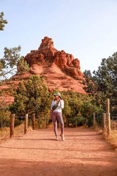 a woman walking down a dirt road in front of a red rock formation and trees