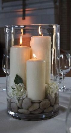 three white candles are sitting in a glass vase with rocks and greenery on the table