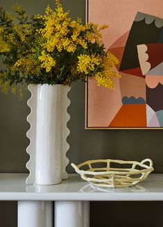 a white vase with yellow flowers in it on top of a table next to a painting