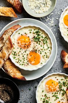 three plates filled with eggs and bread on top of a gray surface next to other dishes