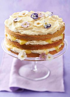 a layered cake with white frosting and purple flowers on top is sitting on a glass plate