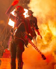 two people dressed in black and red are playing guitar on stage with flames behind them