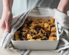 a person holding a dish with bread and blueberries in it on top of a table