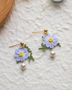 two small blue flowers with green leaves and pearls are on top of a white surface