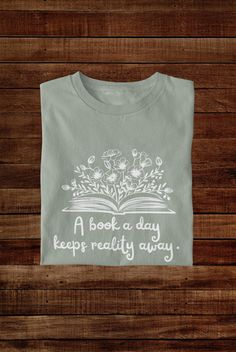 Escape into the pages with our "A Book a Day Keeps Reality Away" t-shirt, perfect for book lovers who prefer fiction over the real world. Made from soft, breathable cotton, this tee combines comfort with a fun, relatable message for avid readers. Whether you're curled up with a new novel or visiting your favorite bookstore, it's the ideal shirt to show off your love for reading. Available in multiple sizes, it also makes a great gift for fellow bookworms who enjoy a daily literary escape! This extra soft tshirt fits like a favorite straight from the box, so don't be surprised when it quickly becomes an actual favorite. The ribbed-knit collars, tapered shoulders, and dual side seams mean this shirt will keep it's shape  and quality over time. A tear-away label minimizes skin irritation.  Co Tshirts For Book Lovers, Bookish T-shirt With Funny Print For School, Reading Tshirt, Bookish T-shirt With Funny Text And Crew Neck, It’s A Good Day To Read A Book Shirt, Cheap Bookish T-shirt With Funny Text, Reading Humor, Avid Reader, Gifts For Bookworms